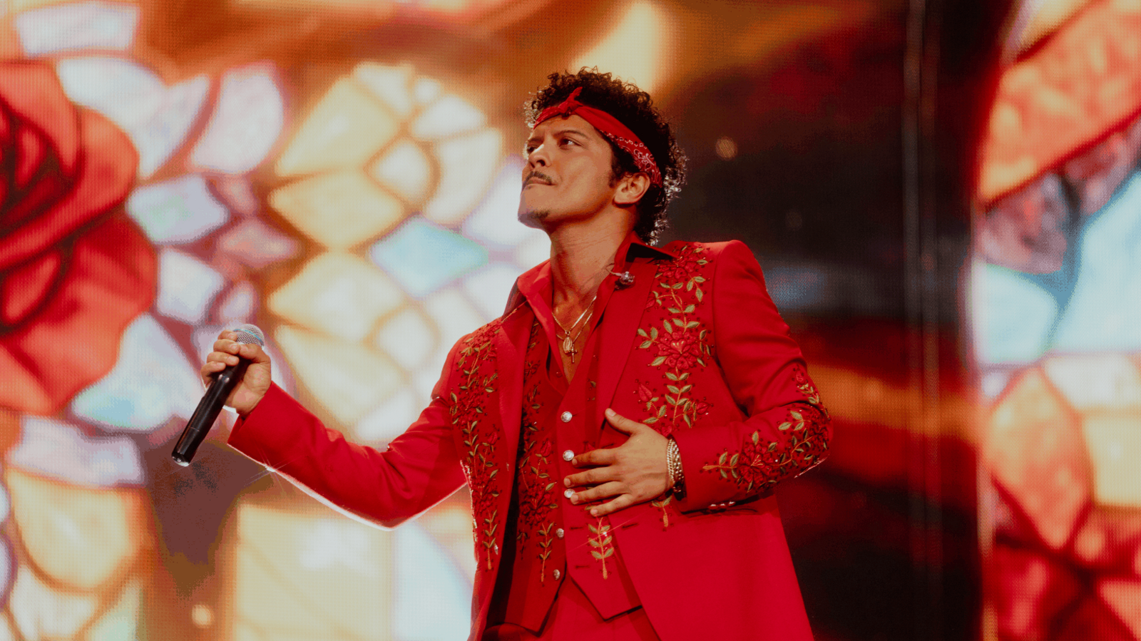 Bruno Mars holds a microphone and performs at Wembley stadium wearing a red suit with gold flower detail. He also wears a red headband.