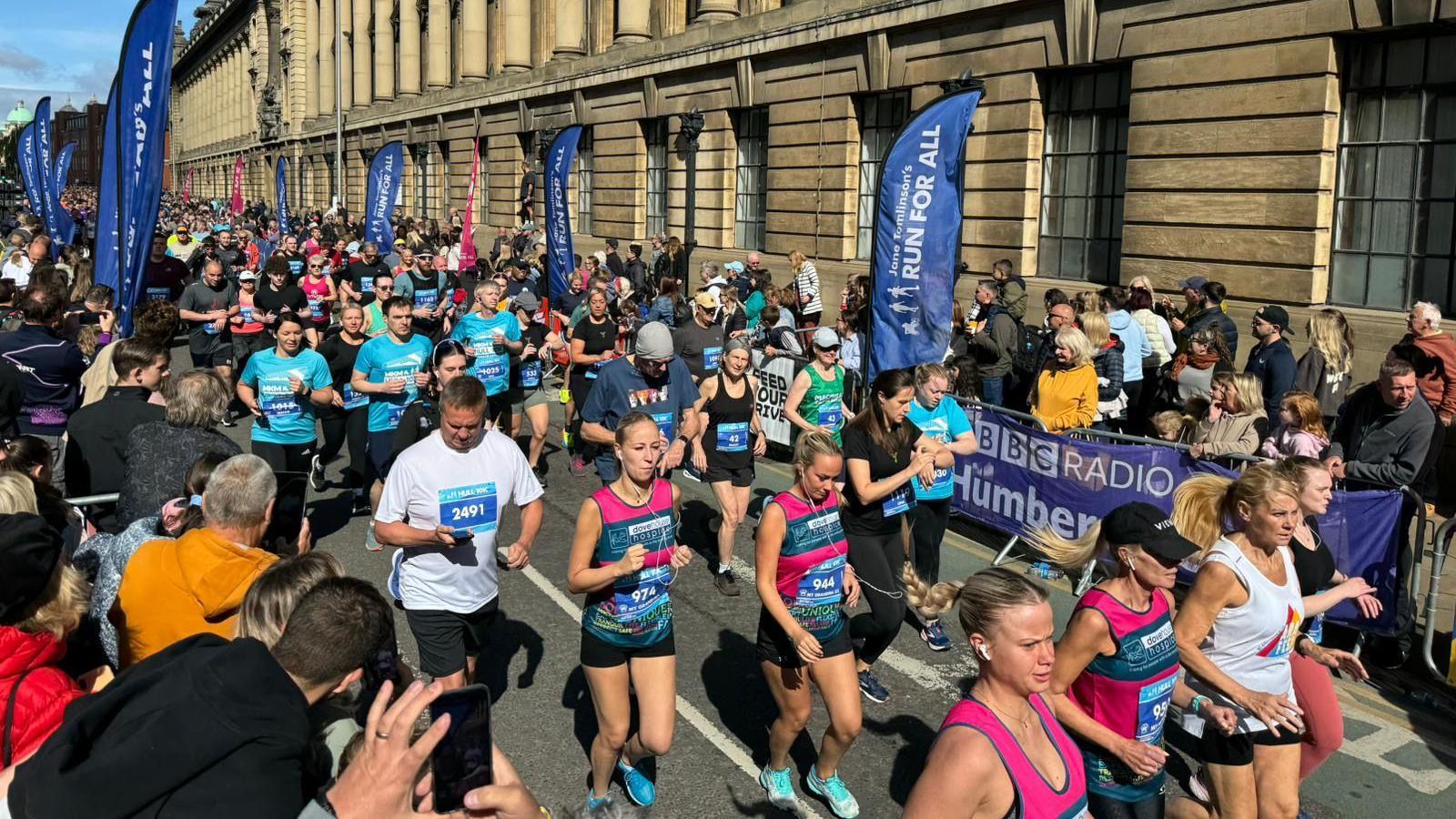 A large group of runners pass the Guildhall in Hull city centre as they take part in the city's 2024 10K race. It is a sunny day and there are large crowds cheering the runners on. There are various banners for the event on display.