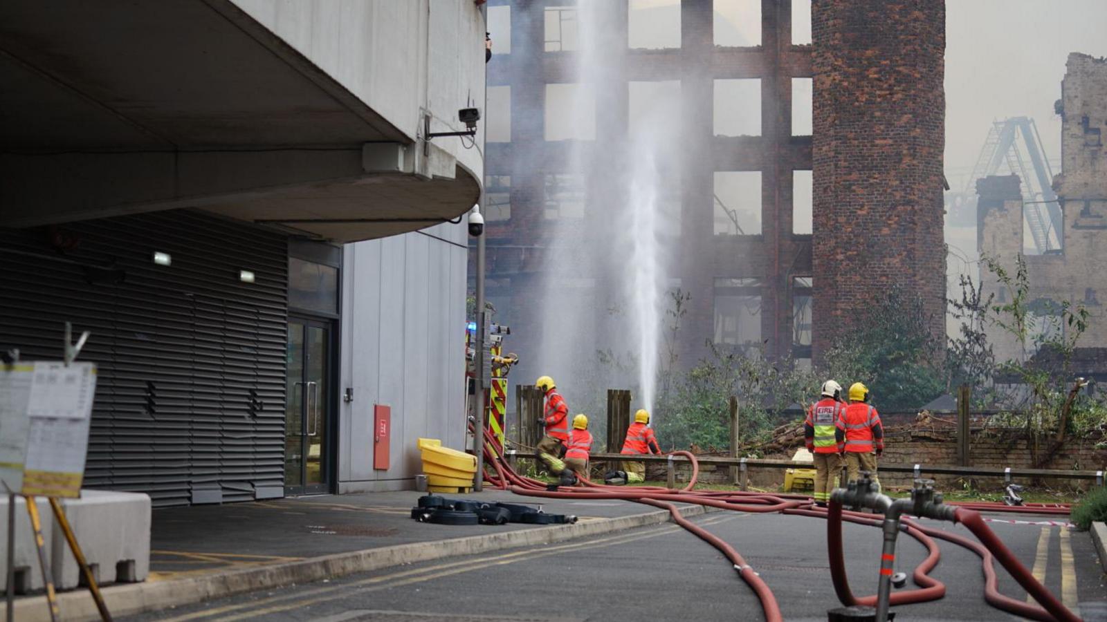 Firefighters wearing yellow helmets and red reflective jackets spray jets of water from hose reels towards a large, smoking red brick building, which has largely aleady been gutted by the fire.