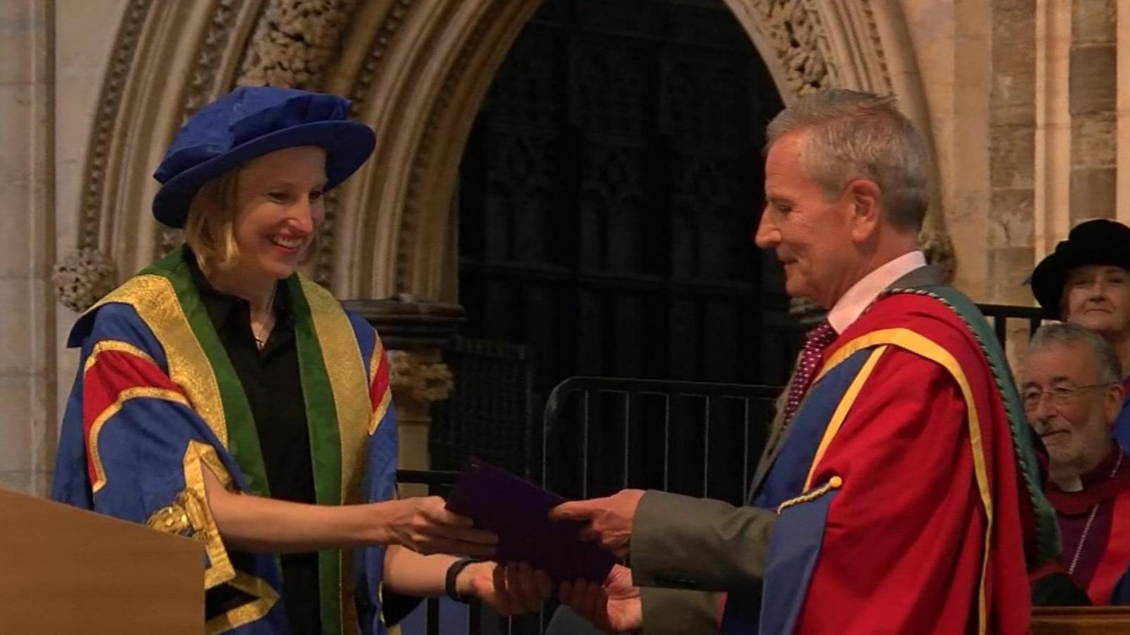 Dr Tracy Borman, wearing a blue hat and the colourful robes of Bishop Grosseteste University presents an honorary doctorate to Peter Levy, who is wearing similar robes over a grey suit, in front of a grand doorway at Lincoln Cathedral.