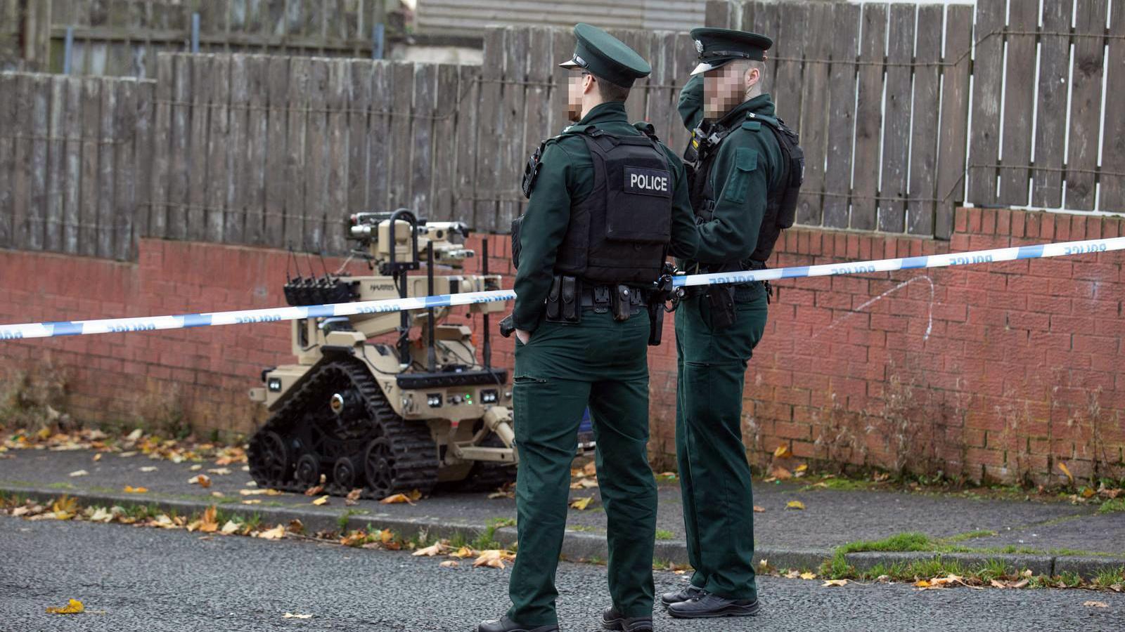 Two male police officers standing at a blue and white police tape. Their faces are blurred and they are wearing a bottle green police uniform with a black bullet proof vest. There is an ATO machine which is a brown colour with black wheels.