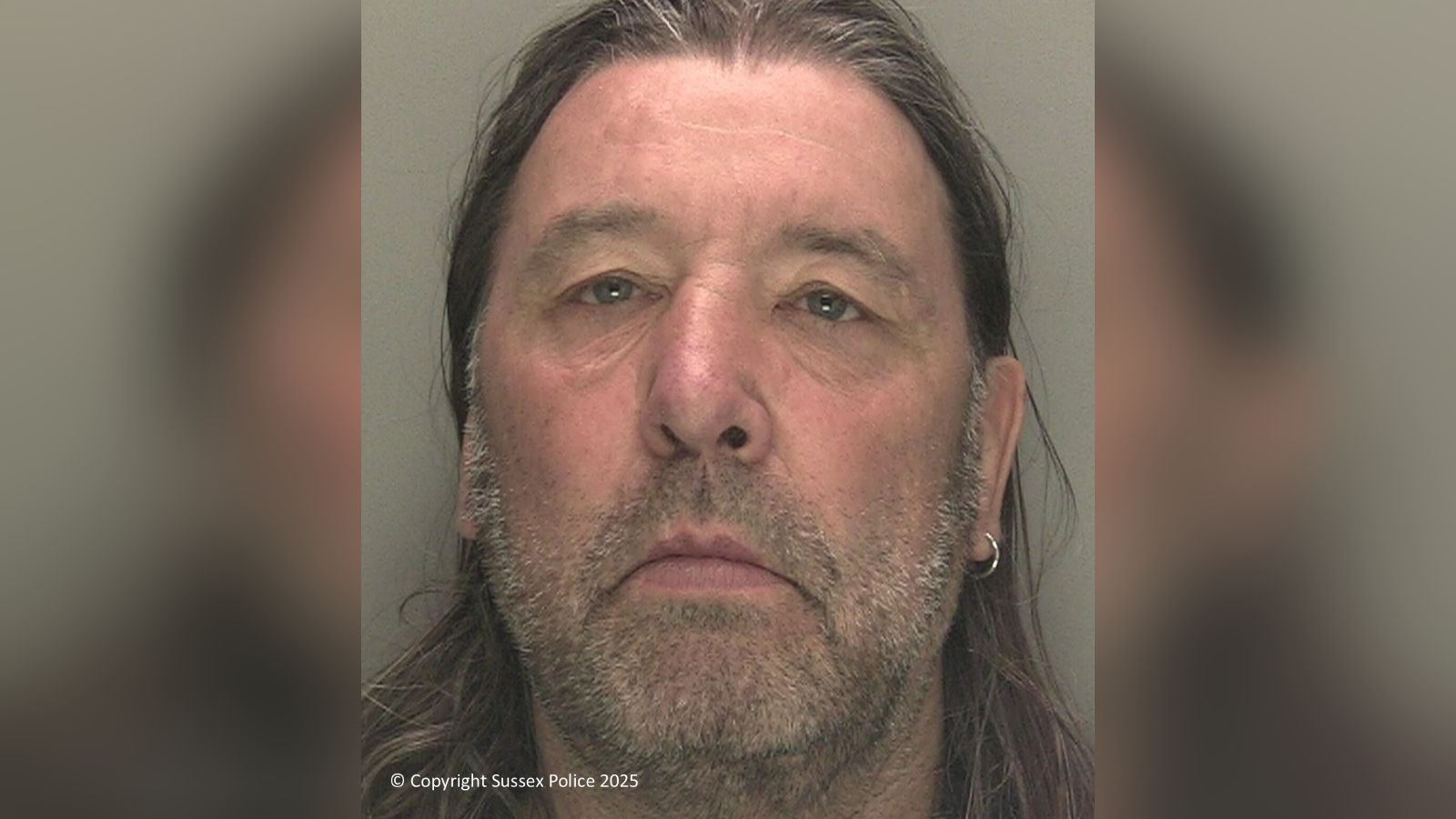 A mugshot of a man with an earring, grey stubble, and long hair.