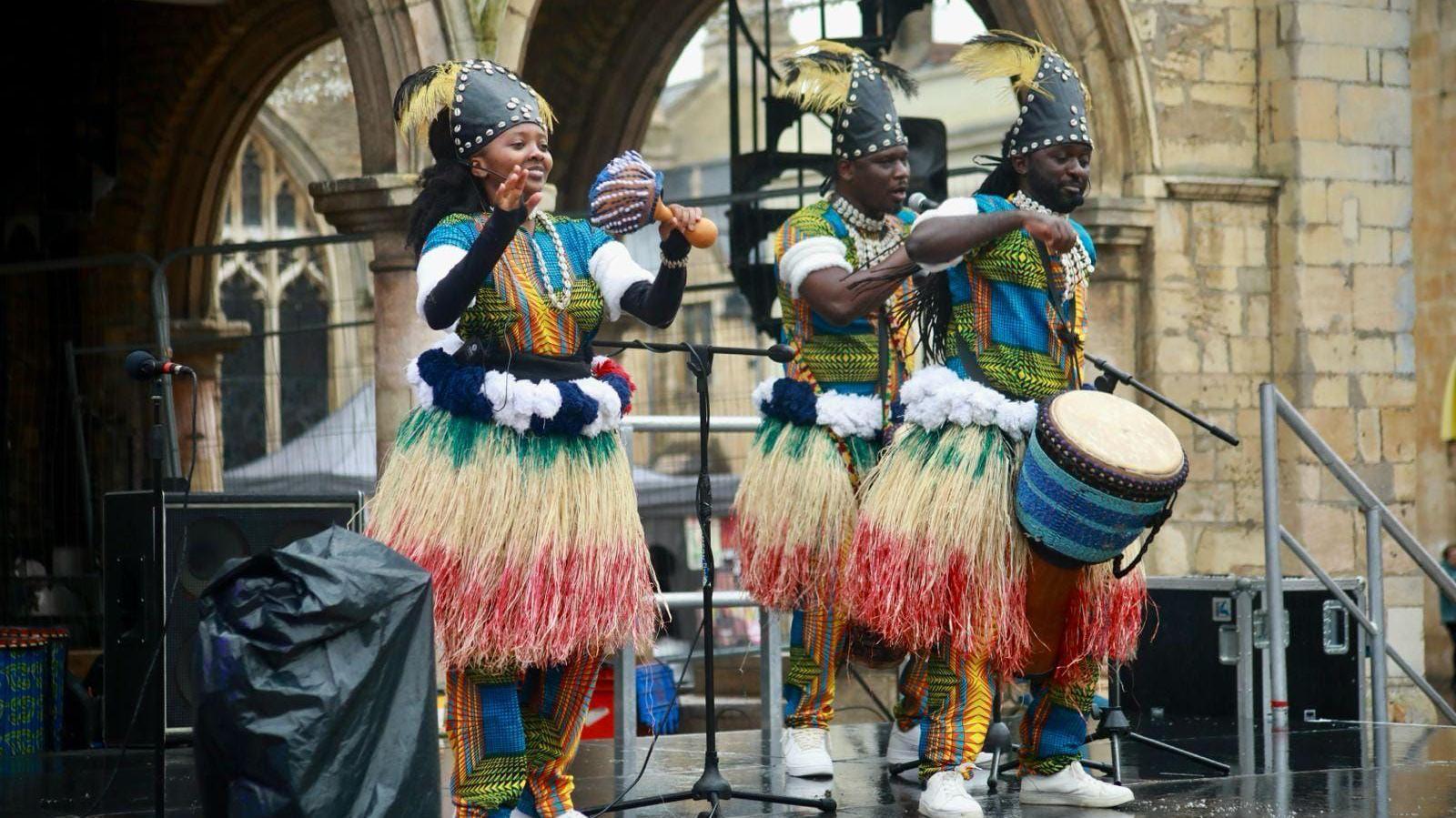 A group of three performers - a woman and two men on a stage - wearing traditional African outfits with straw-looking skirts and playing traditional drums. One is singing into a microphone.