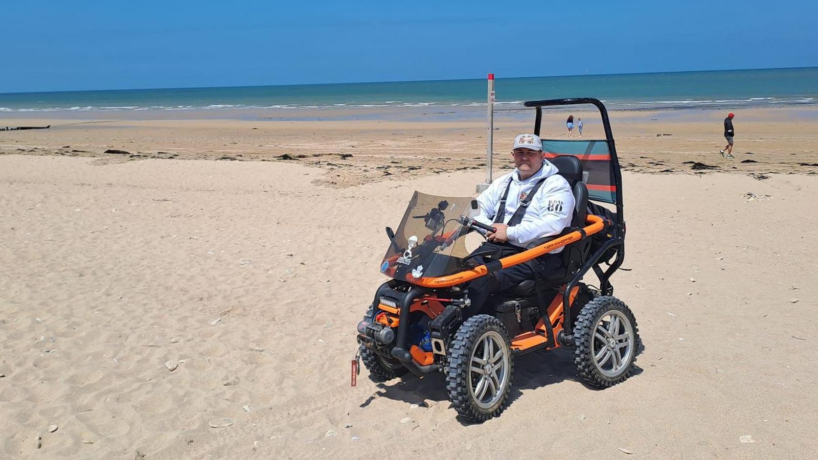 George Pas wearing a grey hoodie and cap to commemorate the 80th anniversary of the D-Day landings. He sitting in his orange off-road wheelchair on Juno beach. It is a sunny day and the tide is out.
