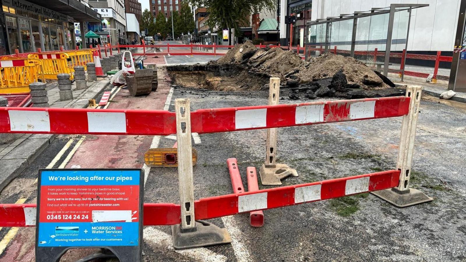 A construction site cordoned off with red and white barriers. A large hole has been excavated in the ground, surrounded by piles of dirt and debris. There is a sign which reads 'we're looking after our pipes'. In the background are more barriers and construction equipment.