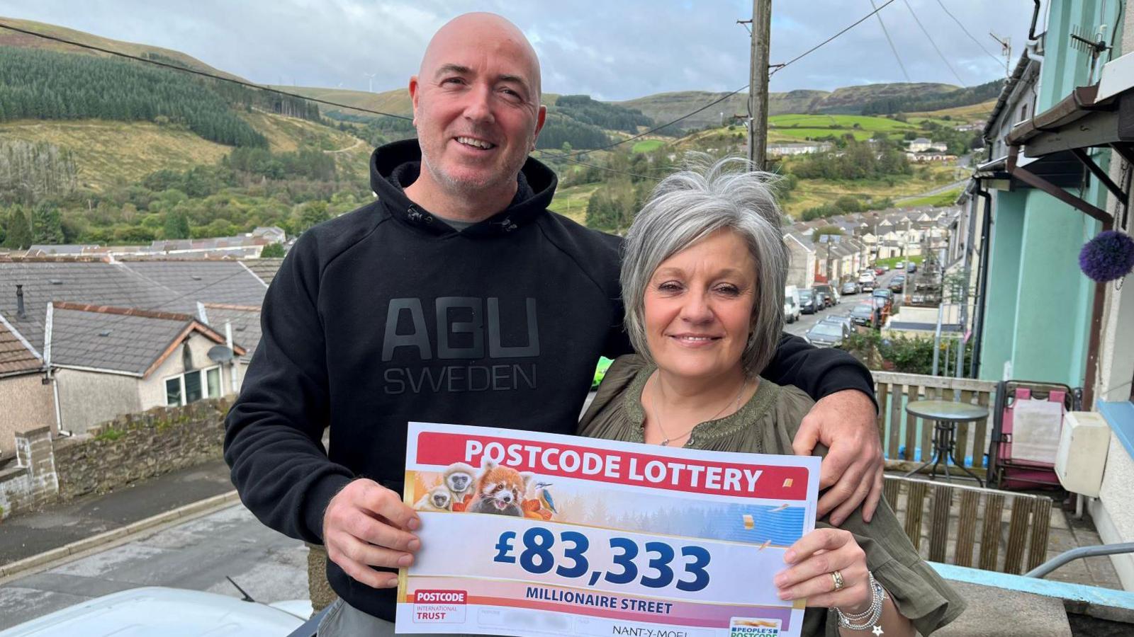 Grant and Katrina Williams on the street holding the winning Postcode Lottery cheque. Grant is wearing a black hoody and has his arm around Katrina. He has a bald head and is smiling at the camera. Katrina has grey hair and is wearing a khaki top, she is also smiling at the camera.