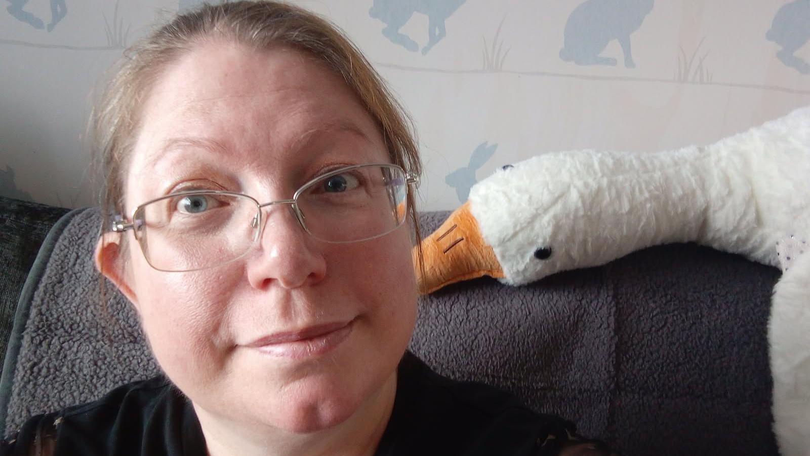 Chrissie Fadipe, 45, has brown hair pulled back tightly over her head, she has glasses on and a black top, she is sitting on a sofa with a toy stuffed goose with his orange beak appearing to nibble her left ear. The wallpaper behind her has a silhouette of grey rabbits.
