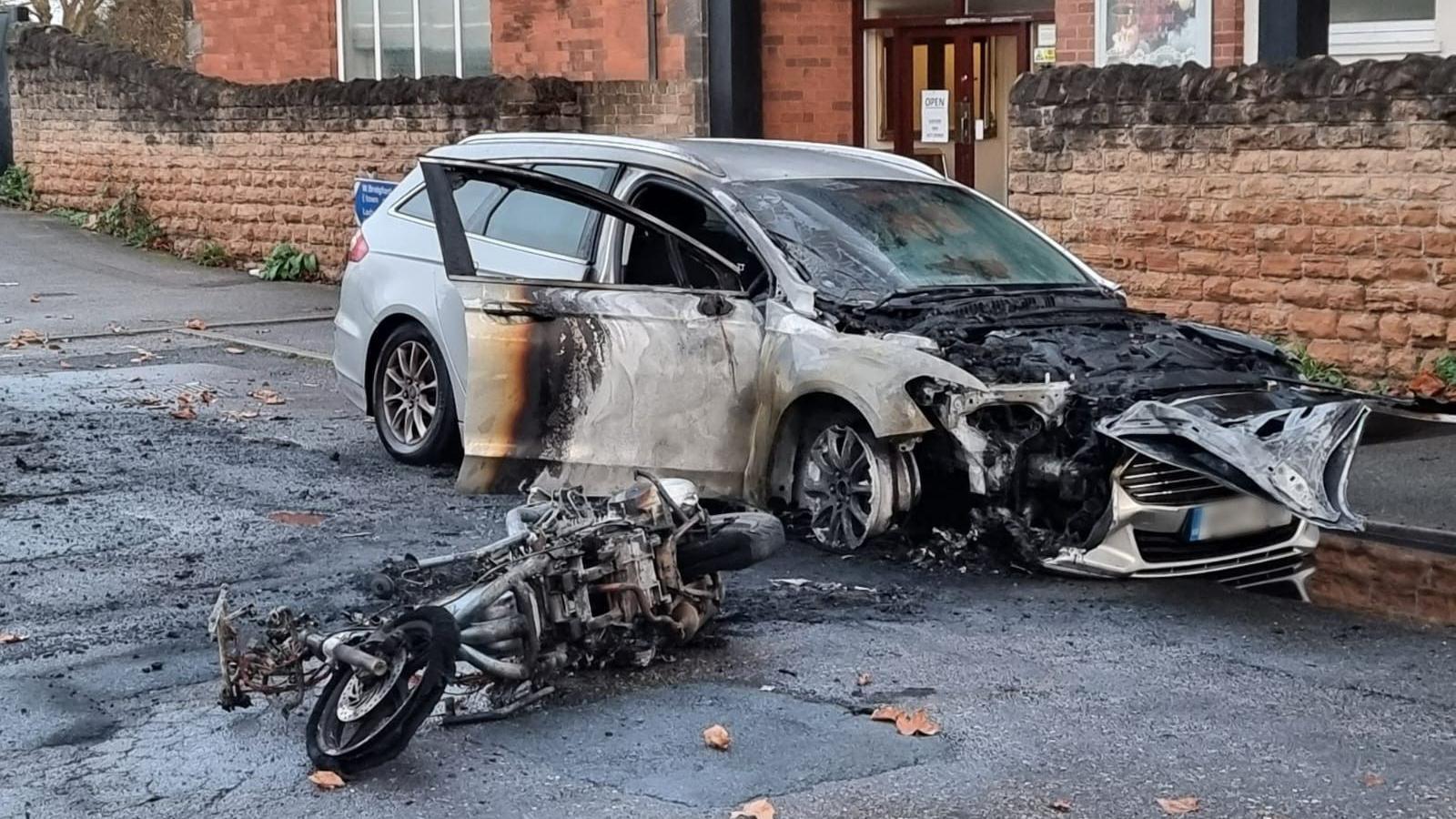 A burnt out parked car and motorbike on its side.