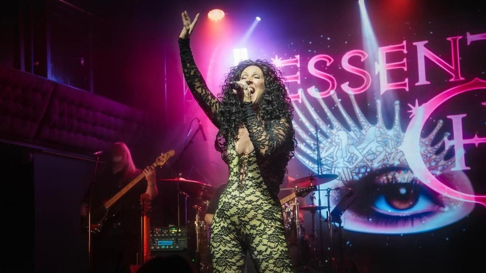 Trisha McCluney dressed as Cher in a lace bodysuit singing on stage, holding a microphone in one hand and with her other hand in the air. Behind her a drummer and a guitar player and behind that a large screen with a picture of an eye.