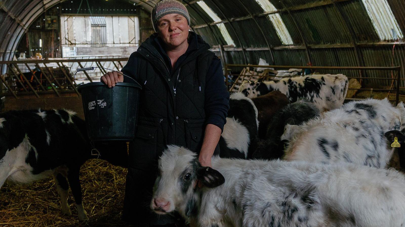 Becky Land holding a bucket in her left hand. Her right hand is placed on a black and white cow which is looking at the camera. There are more cows in the barn behind.