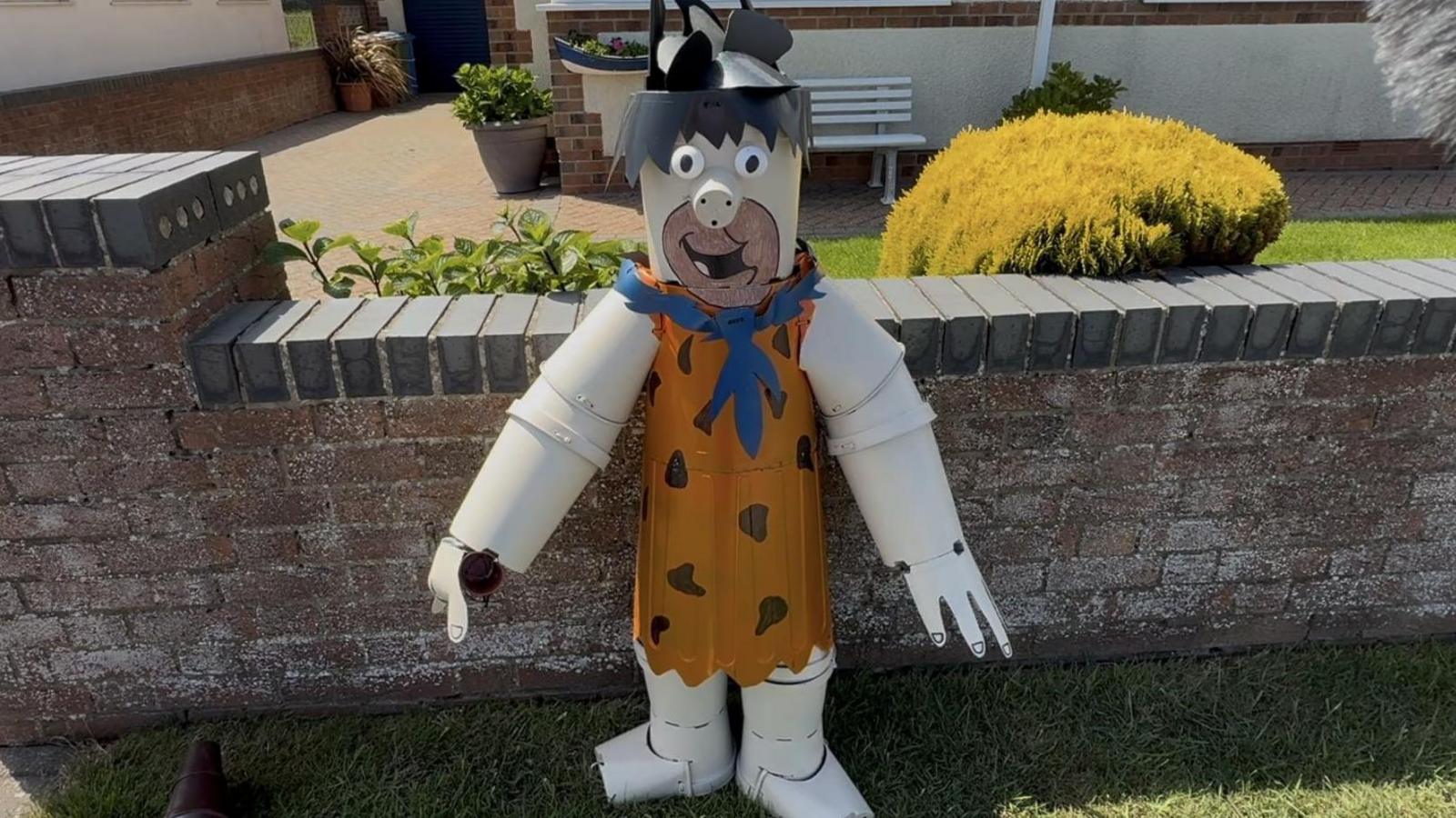 A statue of Fred Flintstone stands in front of a wall. The statue shows the character with black hair and a leopard print outfit.