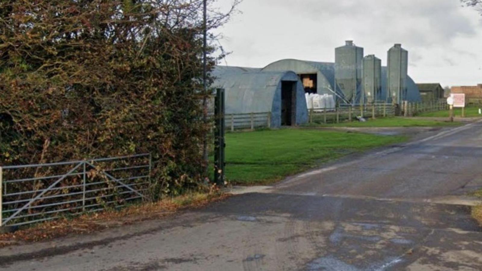 An image of a gate on a road, behind it are several agricultural and industrial buildings.