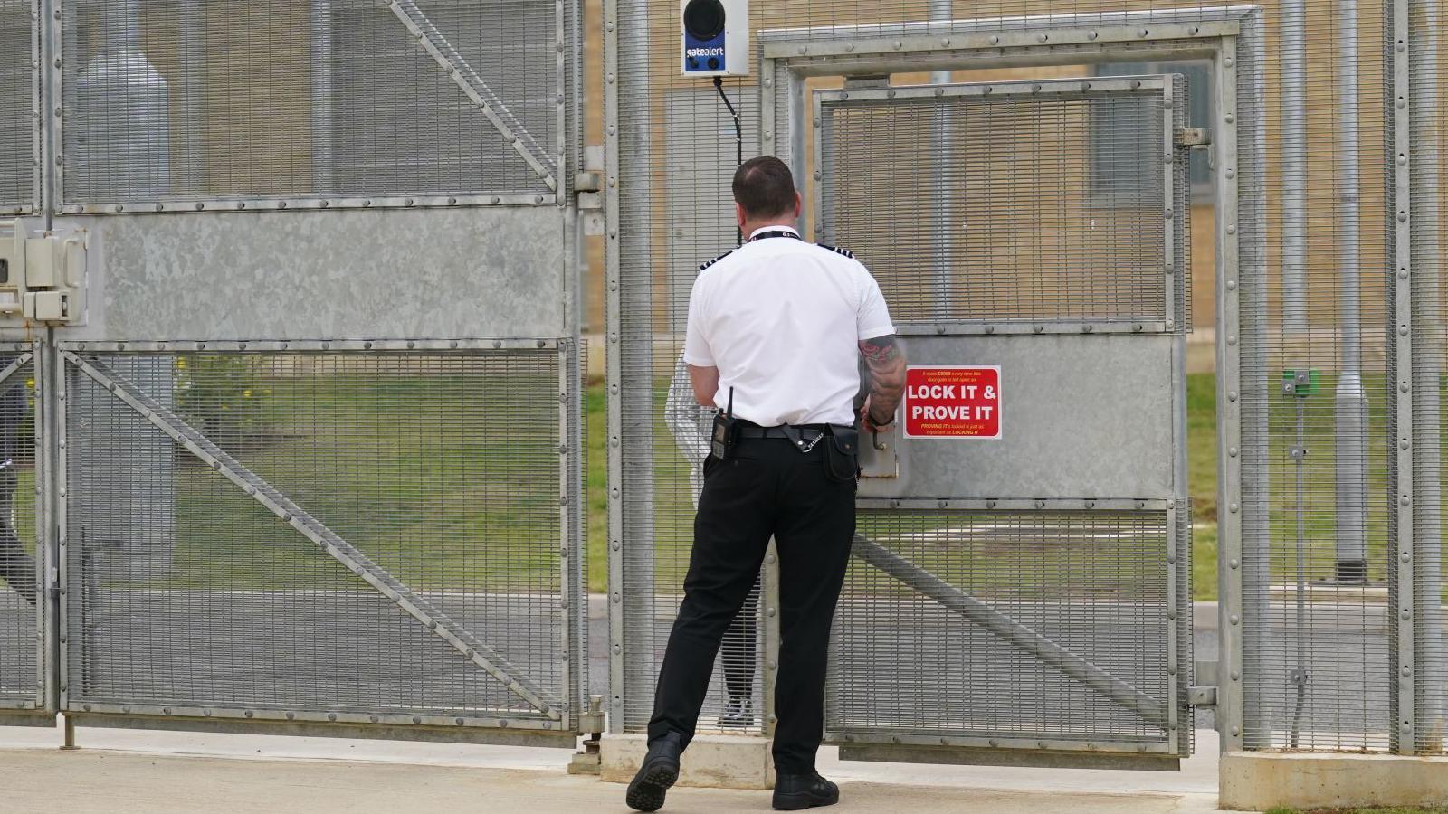 A male prison guard standing outside prison gates. His back is to the camera. He is wearing white and black prison issue uniform. He appears to be talking to another person the other side of the large metal gate and fence.