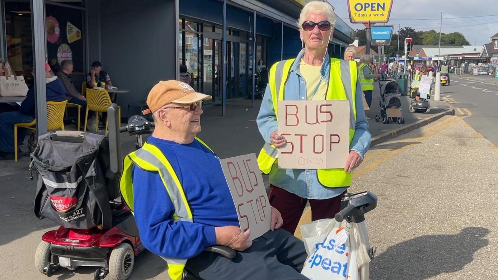 One man, wearing a blue jumper, a bright yellow hi-vis jacket and light orange cap with sunglasses on is sitting in a mobility scooter in a bus lane holding a cardboard sign that reads 'bus stop'. Standing next to him wearing a light blue jacket and also wearing a hi-vis jacket is a woman with white hair and sun glasses holding the same sign. 