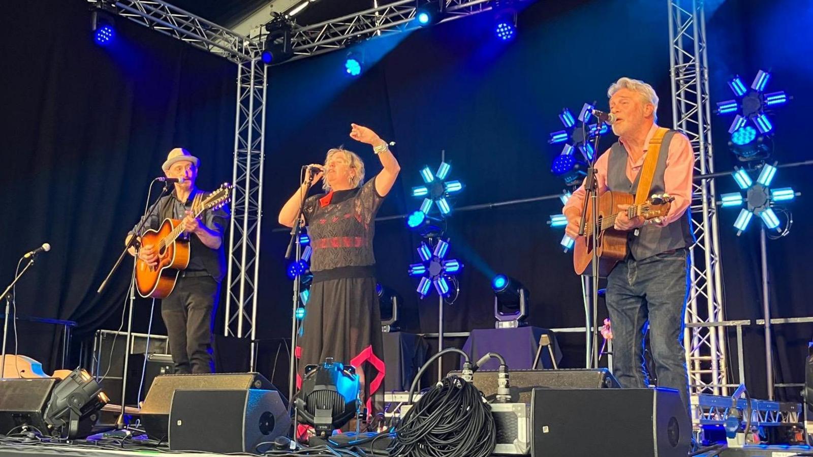 Three people performing on a stage. A woman, who is singing, is in the middle, she is wearing a black top and long, black skirt, she has blonde hair and one hand in the air. A man stands on either side of her, they are playing guitars and also singing. Blue lights in the shape of flowers are lit on a black wall at the back of the stage with amplifiers and lots of wires at the front of the stage. 