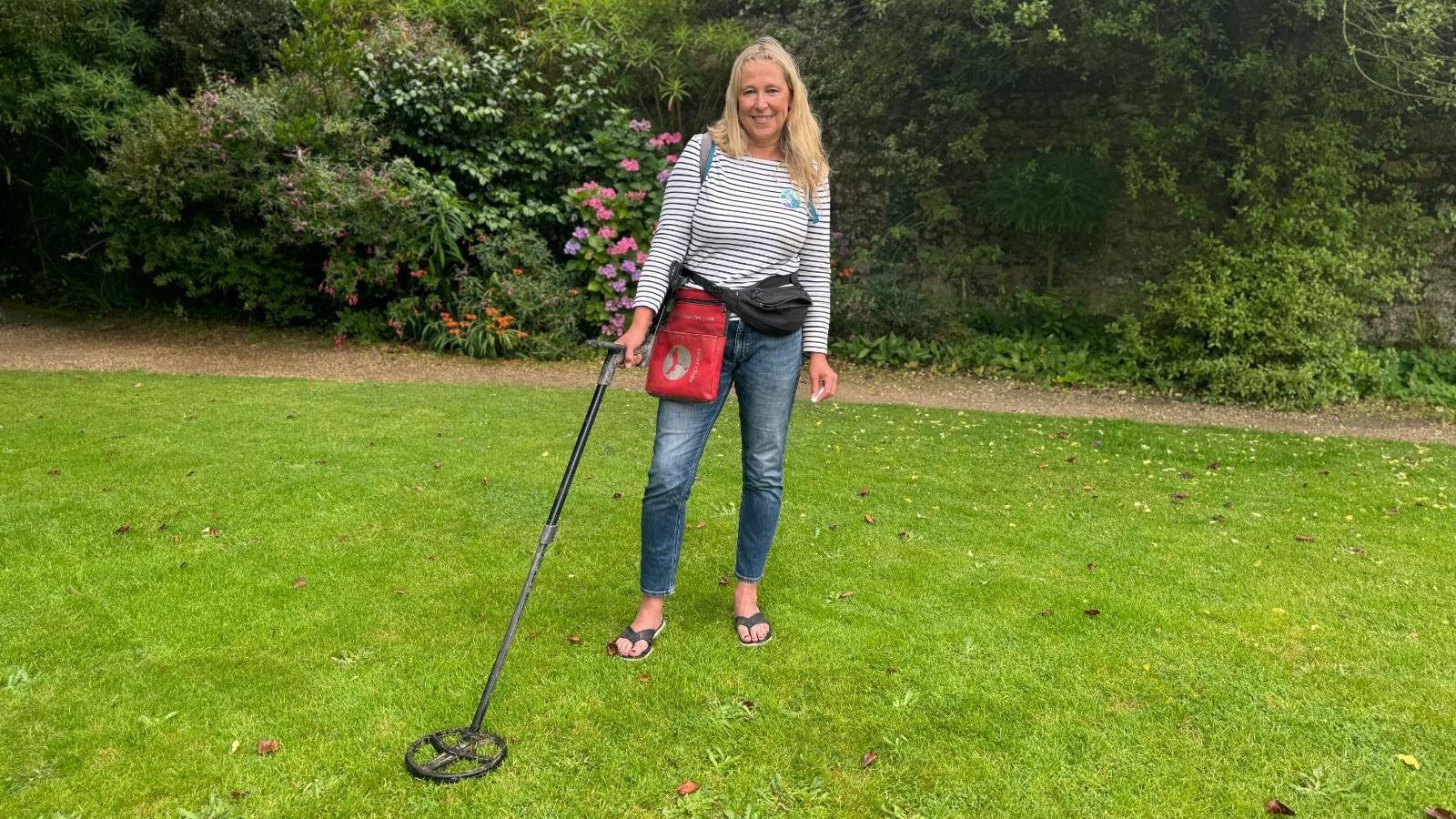 Nicola White - a woman wearing a long sleeved blue and white striped shirt, denim trousers, black flip flops and two bum bags, one red and one black, smiling, blonde hair, holding a metal detector in a green garden with bushes in the background