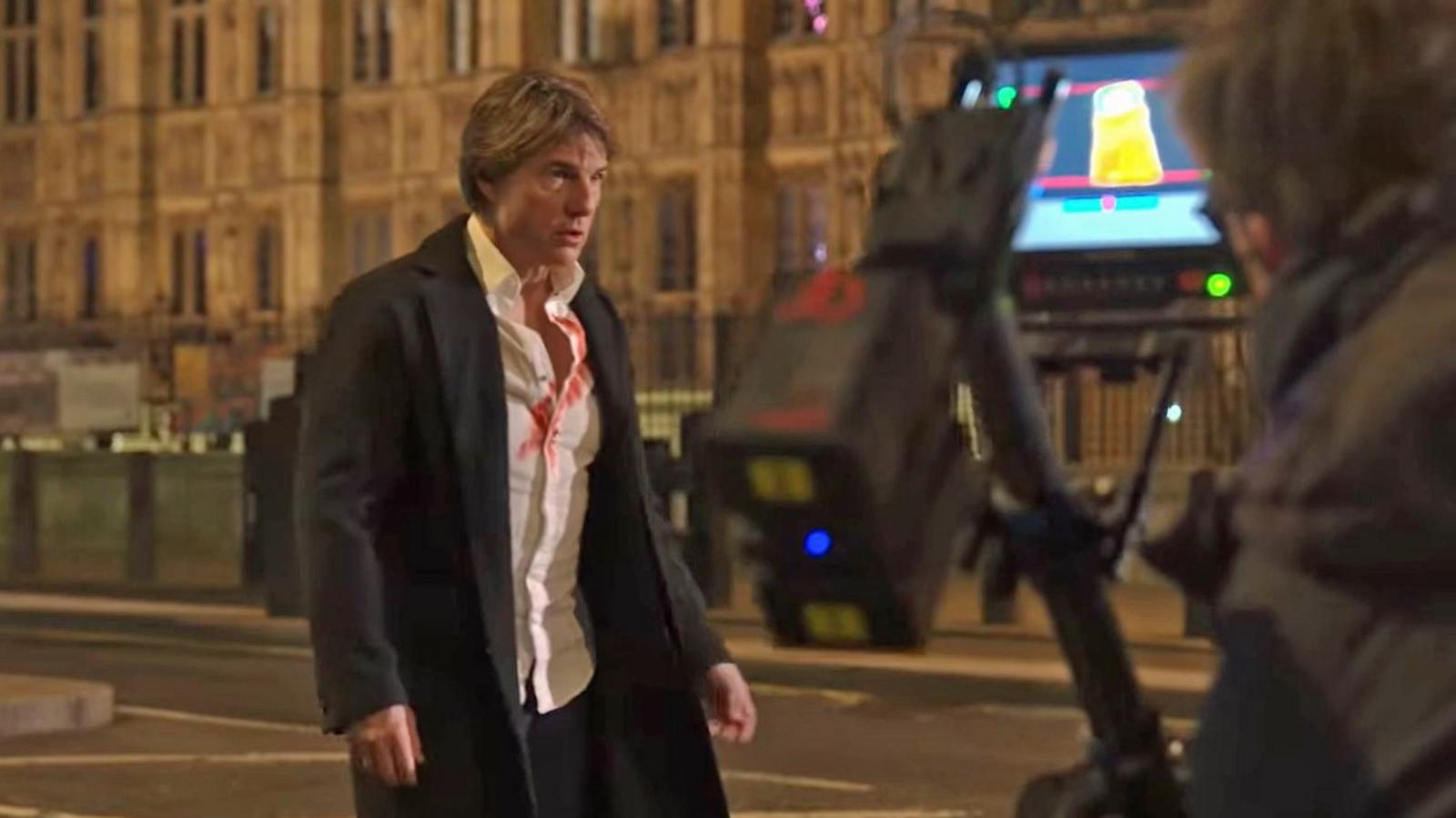 Tom Cruise appears pensive in front of a camera outside the Palace of Westminster. He is wearing a black coat, with a white shirt, slightly open and smeared with red liquid