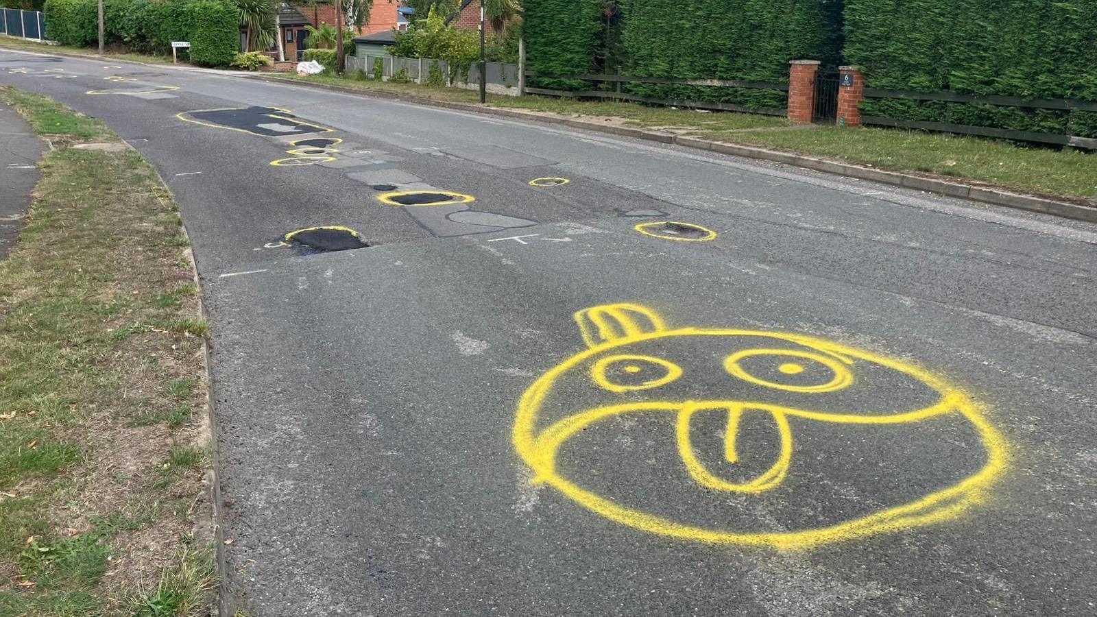 Circles in yellow spray paint around potholes which have been filled in along with a cartoon face with it's tongue out.