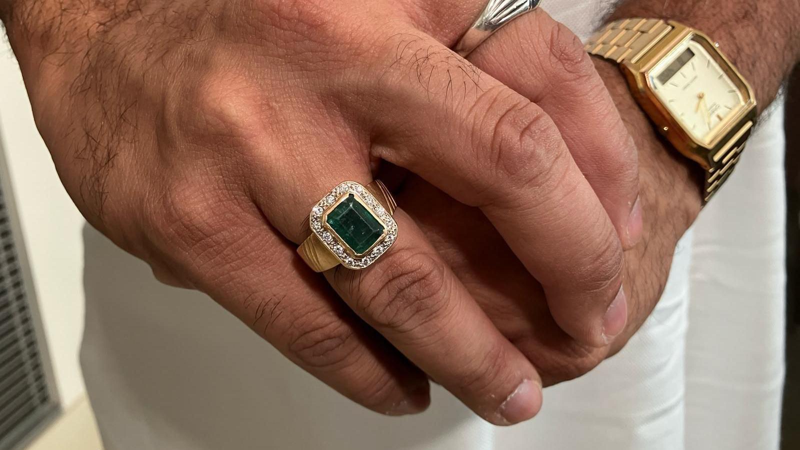 A hand with a gold ring with an emerald in the centre, surrounded by diamonds.