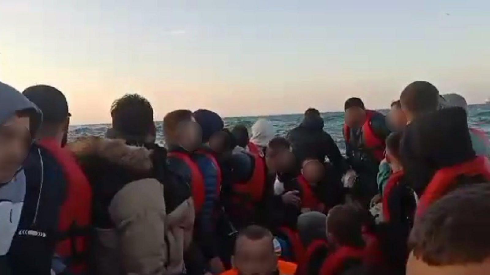 A crowded small boat on The Channel. Men are packed into it as it crosses the water. Their faces have been blurred and they are wearing red life jackets.