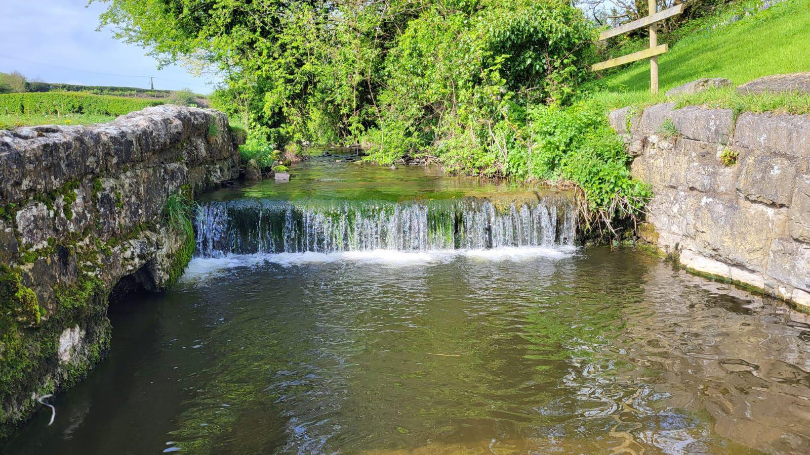 A small weir with water falling down into the river. Stone walls border the river and fields and trees surround it.