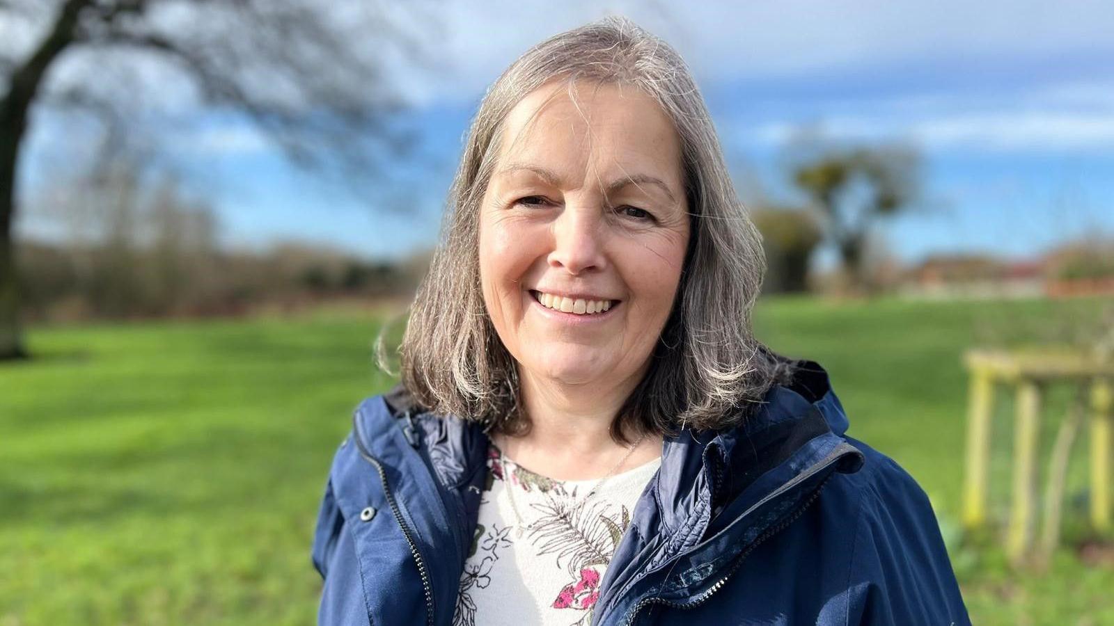 Sarah Murray is standing in a green field. She has shoulder-length grey and brown hair and is smiling at the camera. She has a navy waterproof coat on, with a white floral top underneath.