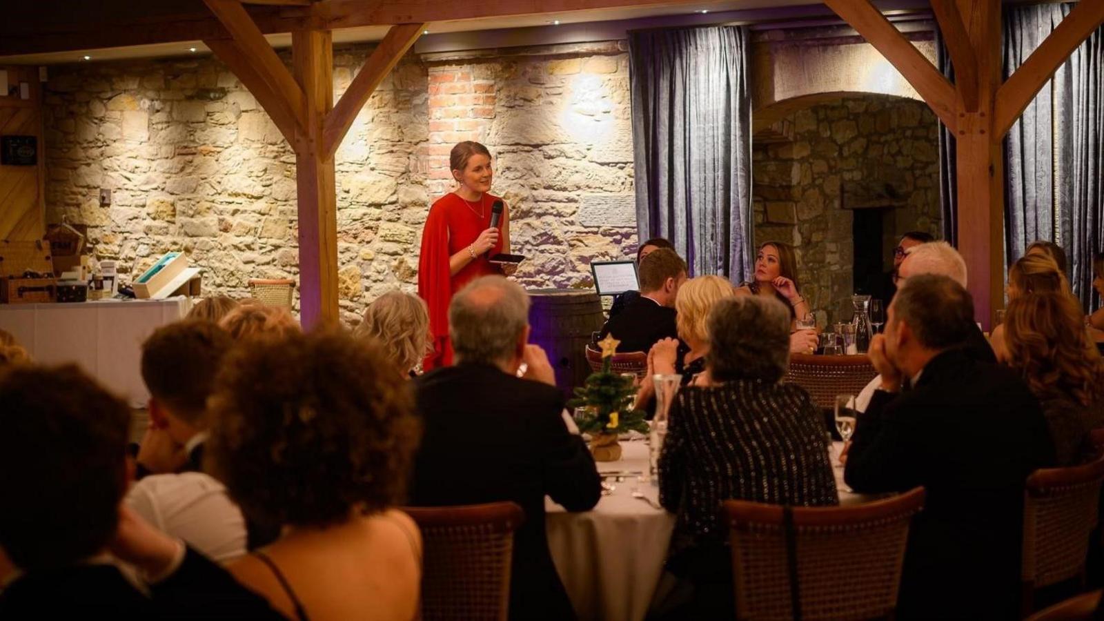 Caitlin Riddell is standing at the front in front of people seated at round tables. She is holding a microphone and is wearing a red dress  