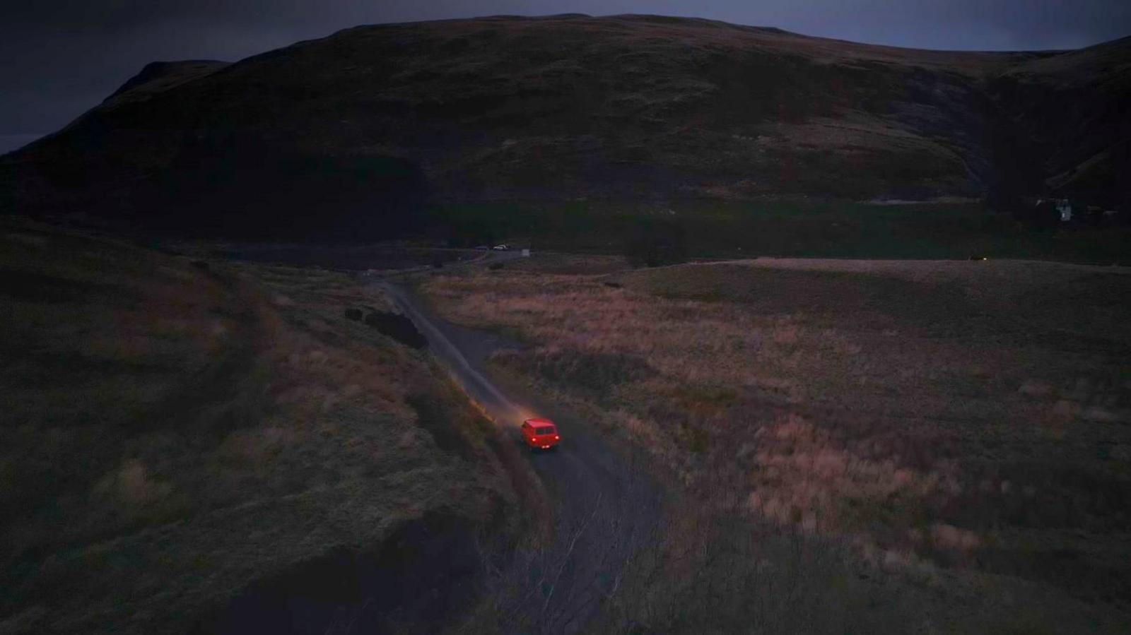 The van is driving up a rural road in the dark. Its lights illuminate the road ahead.