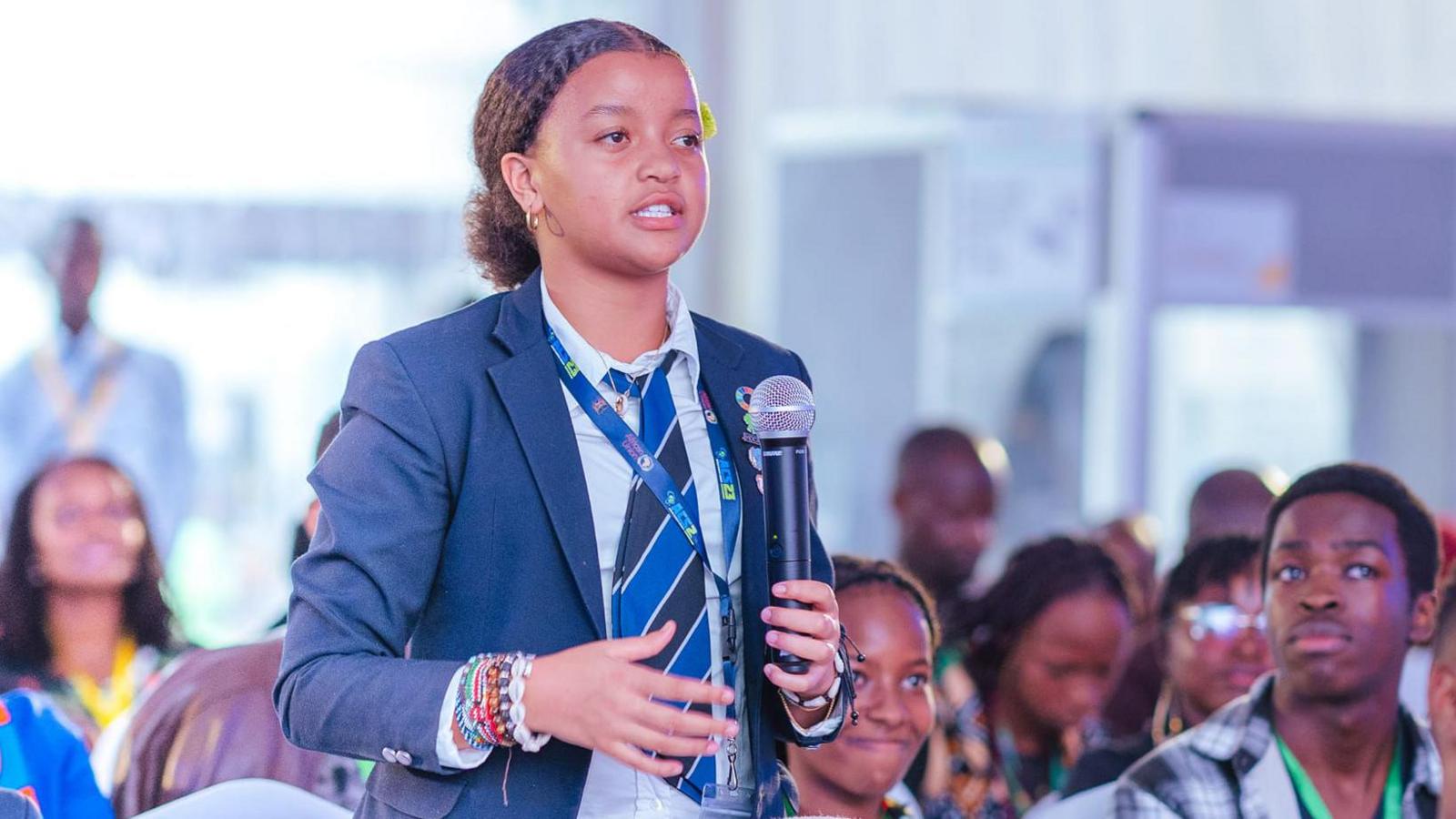 Ellyanne Wanjiku Chlystun, dressed in school uniform, is standing up in an audience of young people and speaks into a microphone.