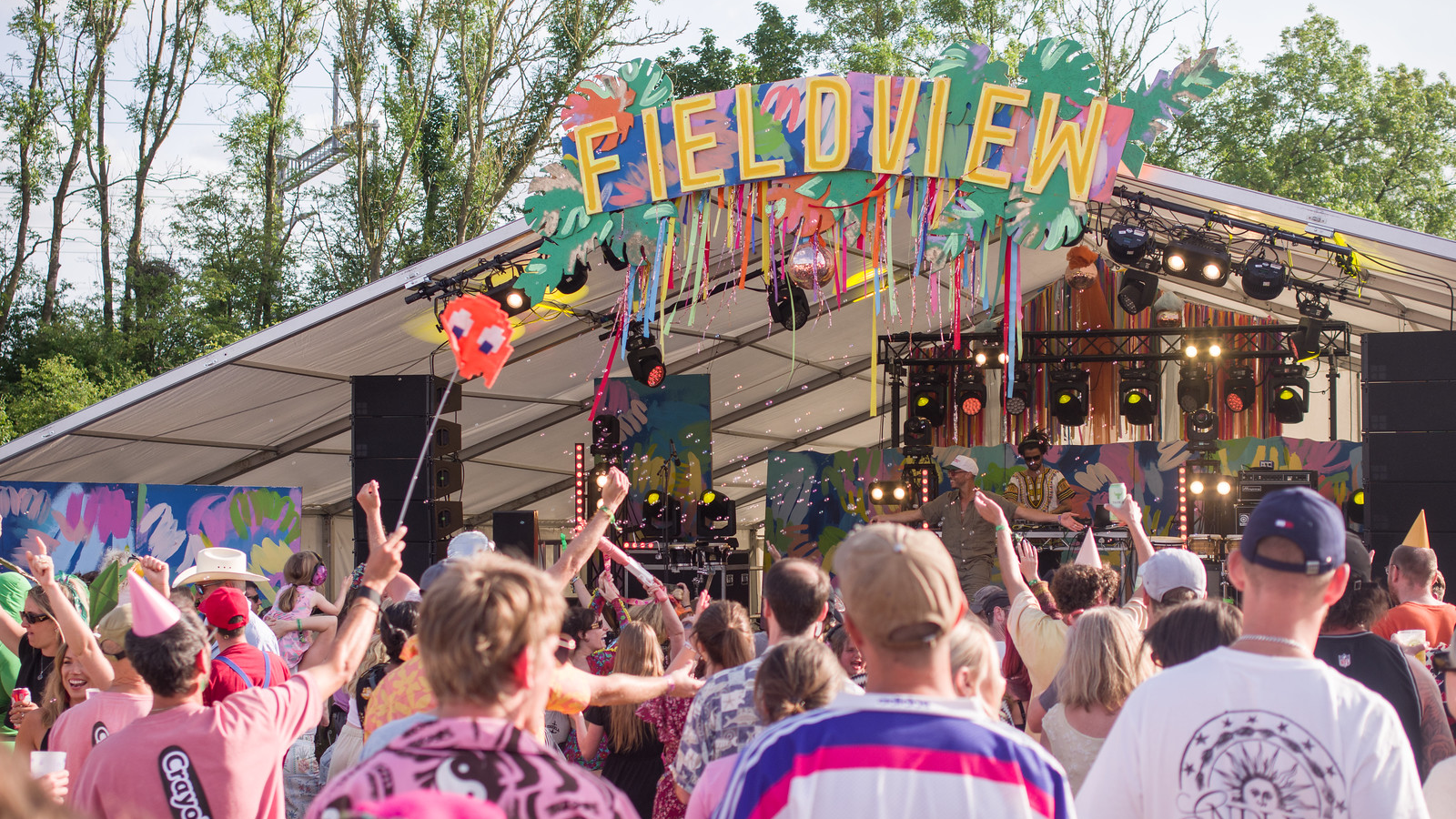 A large group of people stand looking towards an outdoor music stage with a sign reading "Fieldview" on top.
