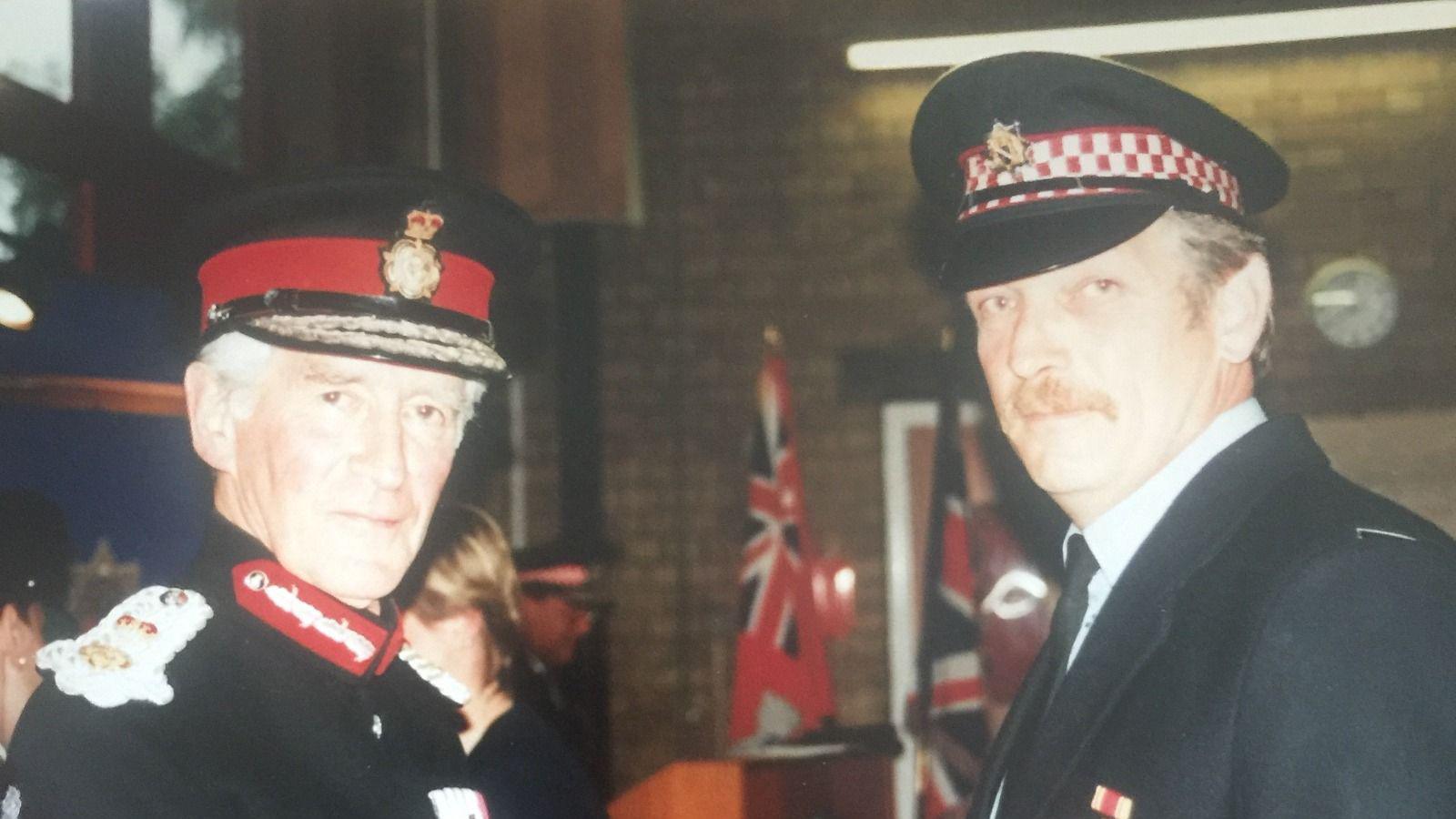 An older man wearing firefighter's dress uniform and a younger man wearing a jacket, tie and firefighter's cap