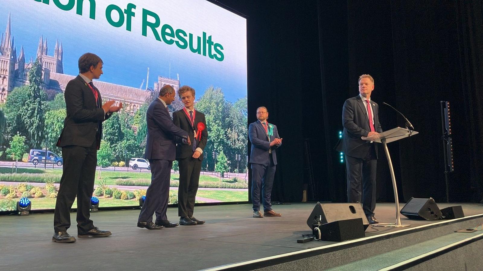 Sam Carling stands centre on the stage and shakes the hand of Conservative candidate Shailesh Vara while other candidates applaud.