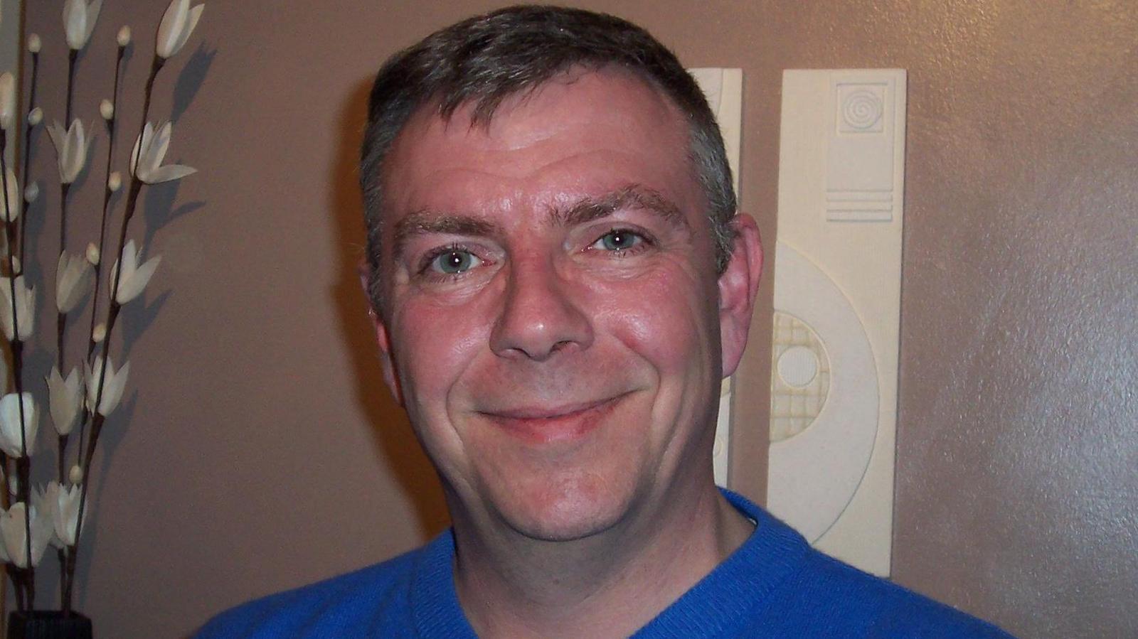 Brian Place, who has grey, short hair, smiling at the camera. He's wearing a blue t-shirt.