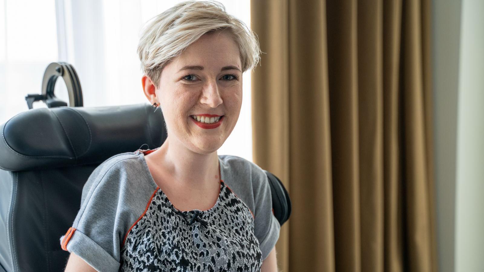 Sarah Rennie has short blonde hair and is sitting in her wheelchair, smiling at the camera. She is wearing a grey leopard print t-shirt and red lipstick