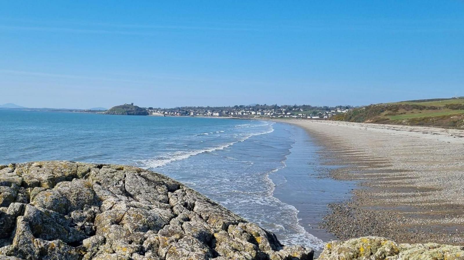 photograph of a rock and beach looking out towards the town of Cricceith with the castle just visible on the rock in the distance. Blue skies above.