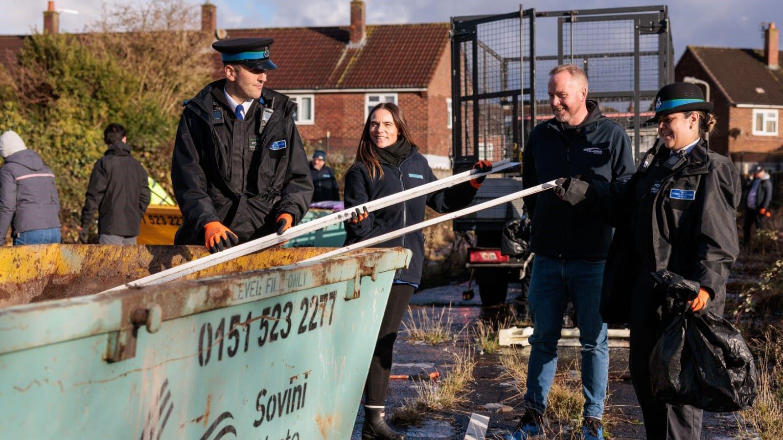 There are two members of the police and two others who are putting building material in a blue skip. They are looking at eachother and smiling