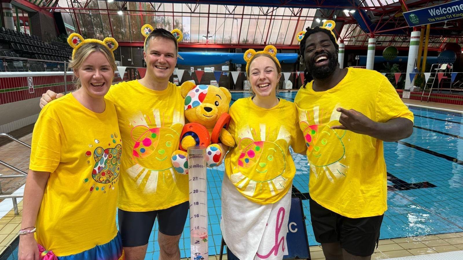 Four BBC Humberside colleagues smiling for the camera in front of a pool. Four of them are all wearing bright yellow tops with Pudsey's face on along with Pudsey ears and a Pudsey teddy in the middle. 