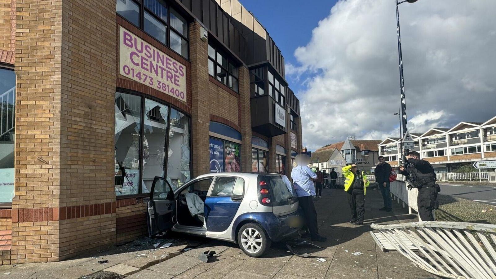 A navy Smart car crashed into a brick building with police officers and onlookers surrounding the scene