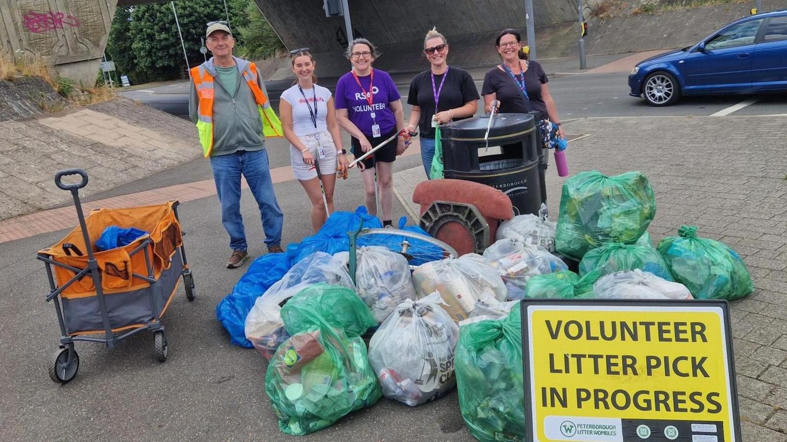 Five people - one male and four female are standing near an underpass. There are numerous bags of collected rubbish on the road in front of them and a yellow sign that says "volunteer litter pick in progress"