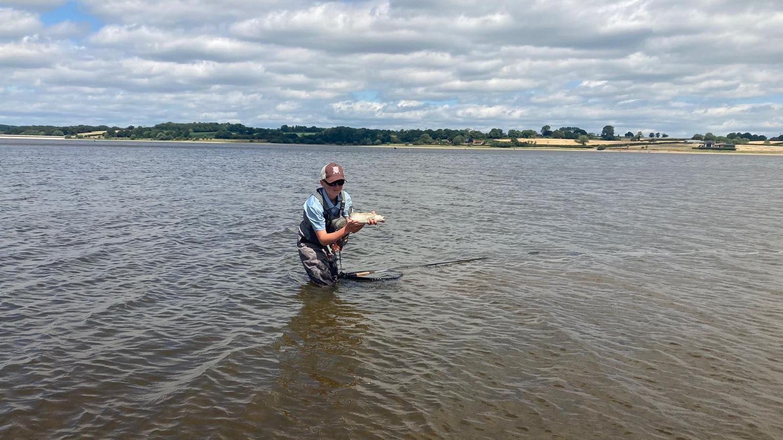 A teenage boy standing in a body of water holding a fish. He has grey waders on and a blue hoodie with a maroon and white cap.