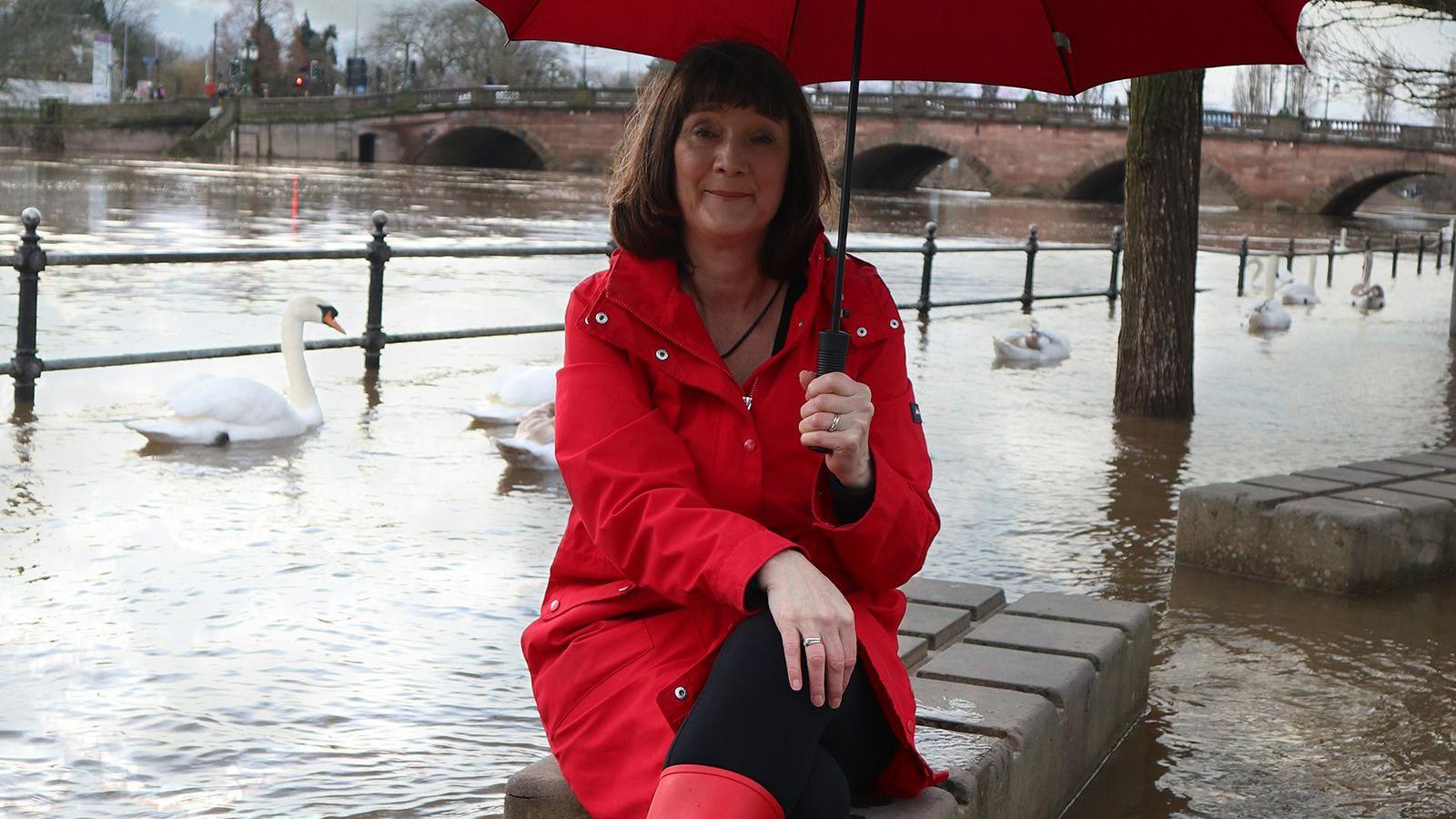 Mary Long-Dhonau sits on a wall by a flooded river with swans behind her. She is wearing a red coat and red boots and is holding a red umbrella. There is a bridge with arches behind her, crossing the river.