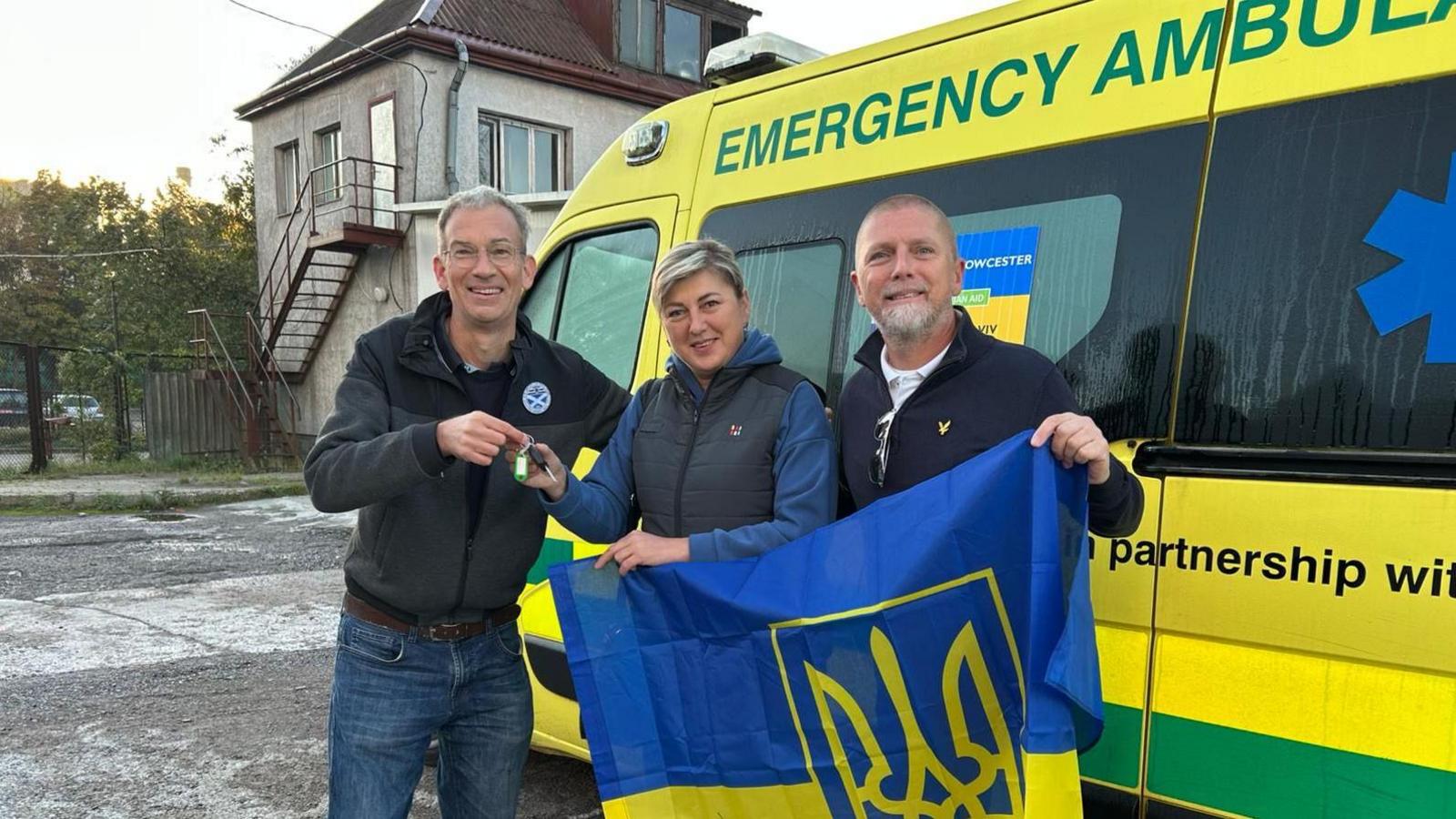 Alex with short dark hair wearing a dark coat, Tetiana with short dark and blond hair wearing a dark blue gilet over a light blue top and Mike with short grey hair and beard, wearing a blue jacket.  Theyare holding a Ukraine flag and standing next to an ambulance.