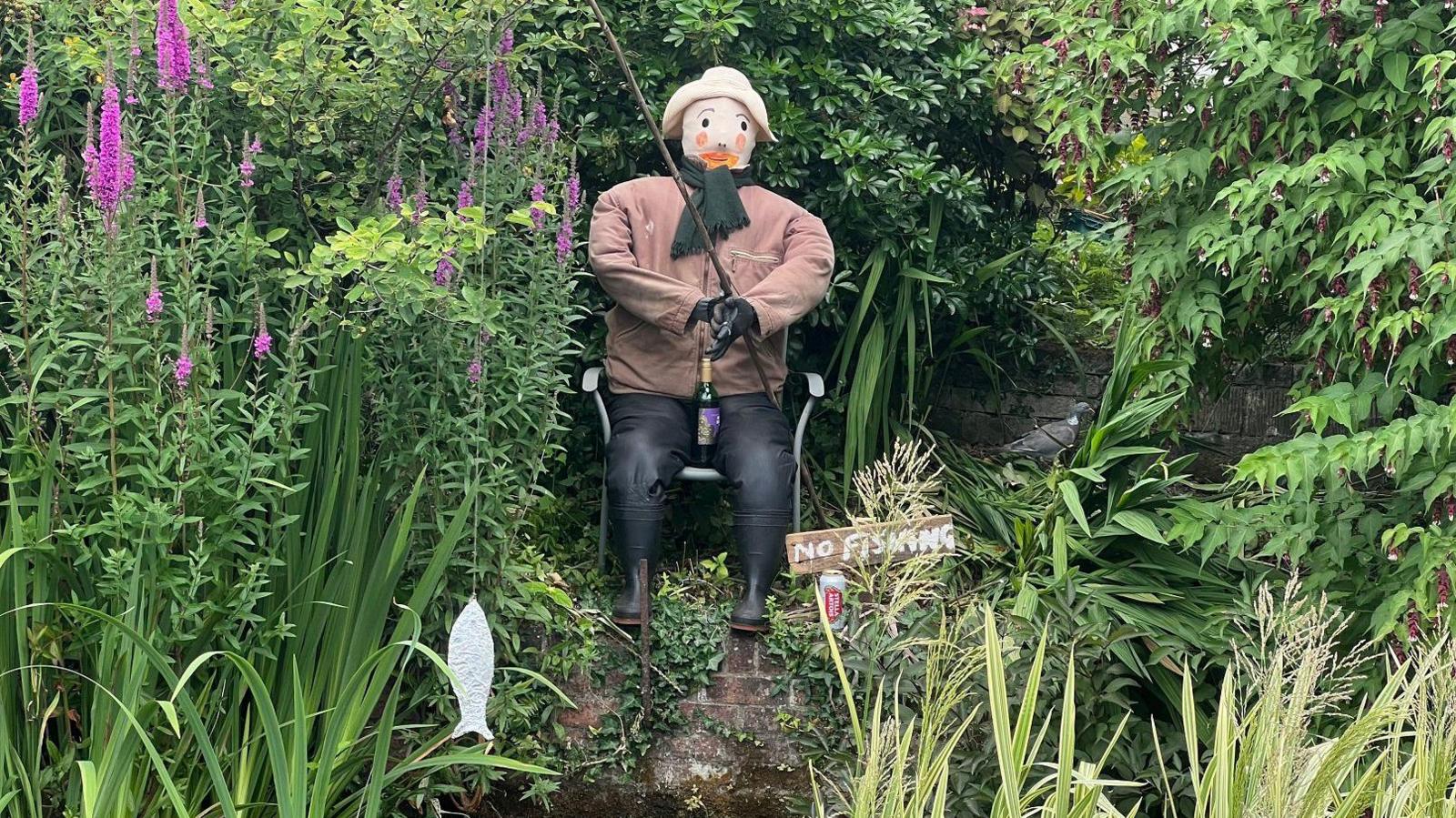 A scarecrow fishes in a pond, ignoring a "no fishing" sign with a can of Stella beside him