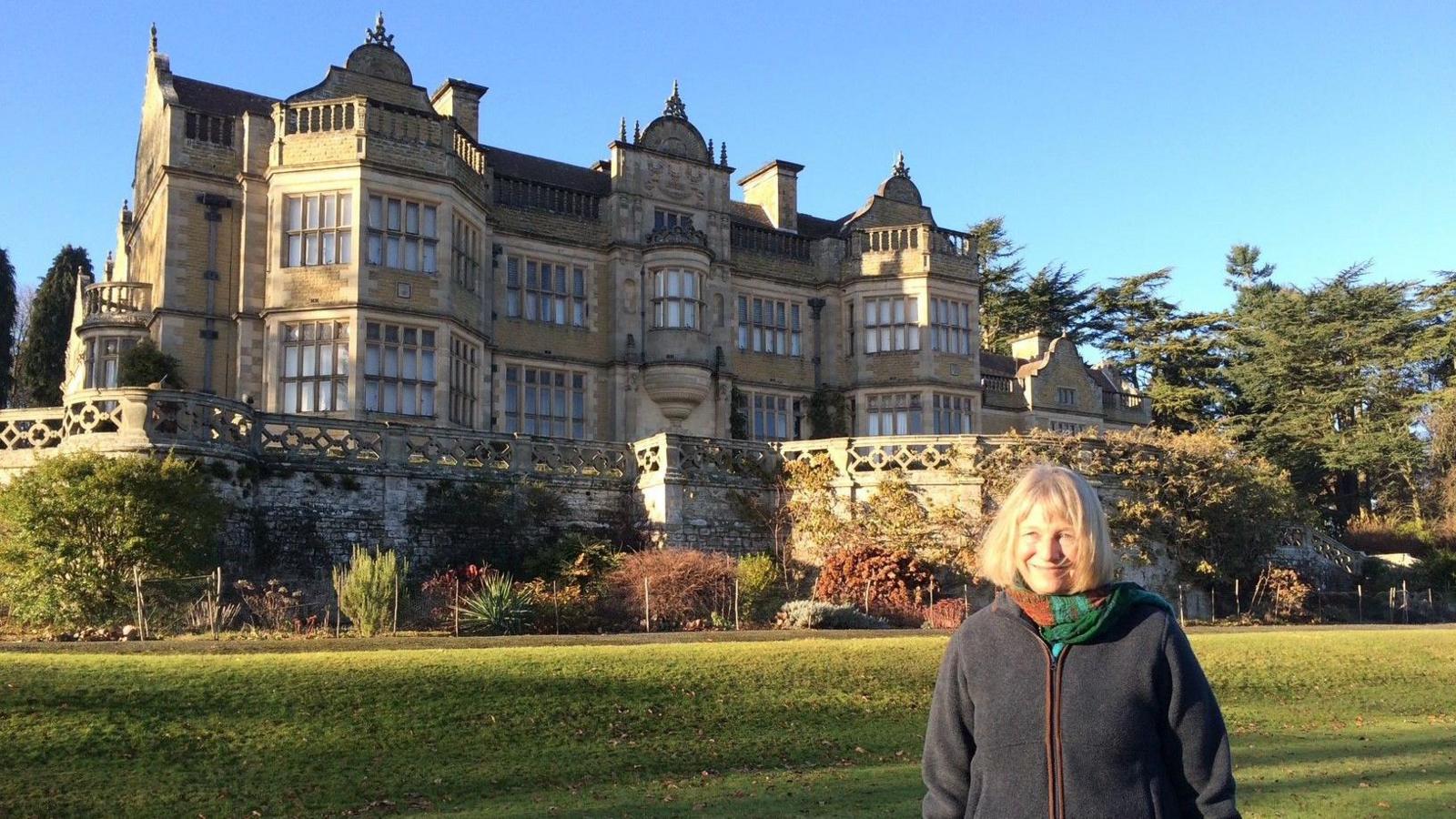 A woman with blonde hair and a dark blue coat standing on a large lawn in front of a large, two-storey house with ornate stonework