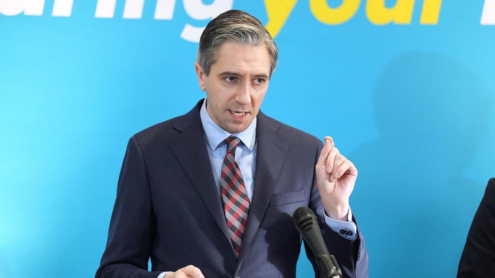 Simon Harris, with greying dark hair, wearing in a dark suit, with his hand raised to emphasise a point while speaking at a podium