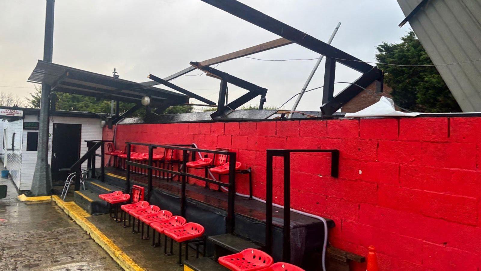 The seating area at Sawbridgeworth Town, with part of the roof missing. You can see the metal beams bent out of shape and a corrugated roof on its side. There are red plastic seats without backs, on two rows - one behind the other. There is a bright red painted wall behind the seats. Te ground looks very wet.