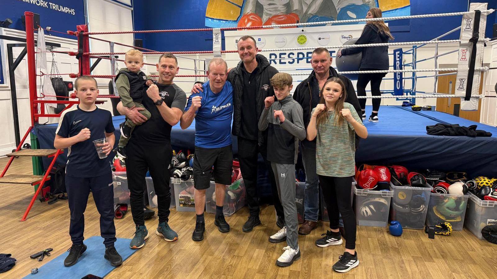 A group of youngsters with four older men holding up their fists and standing next to a boxing ring. They are all wearing trainers and sporting wear.