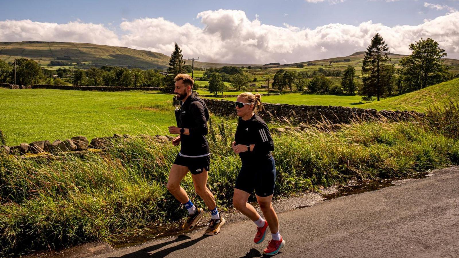 Dr Meg and a running partner run along a country lane with green fields, trees and hills in the background.