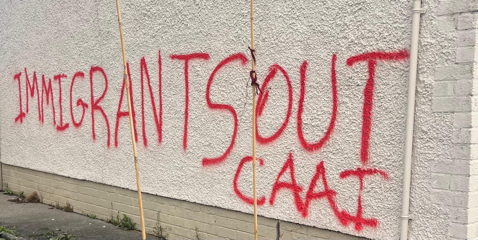 A white gable wall with the words: 'Immigrants out' sprawled in red paint.
A section of the message has been blurred.
A pink rose bush with some flowers can be seen at the side.
