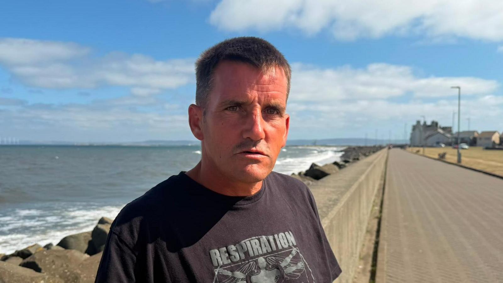 Davey Short has short-cropped brown hair. He is wearing a black t-shirt and he looks like he has been in the sun a lot with slightly red skin. He is standing on a promenade with the sea behind him.